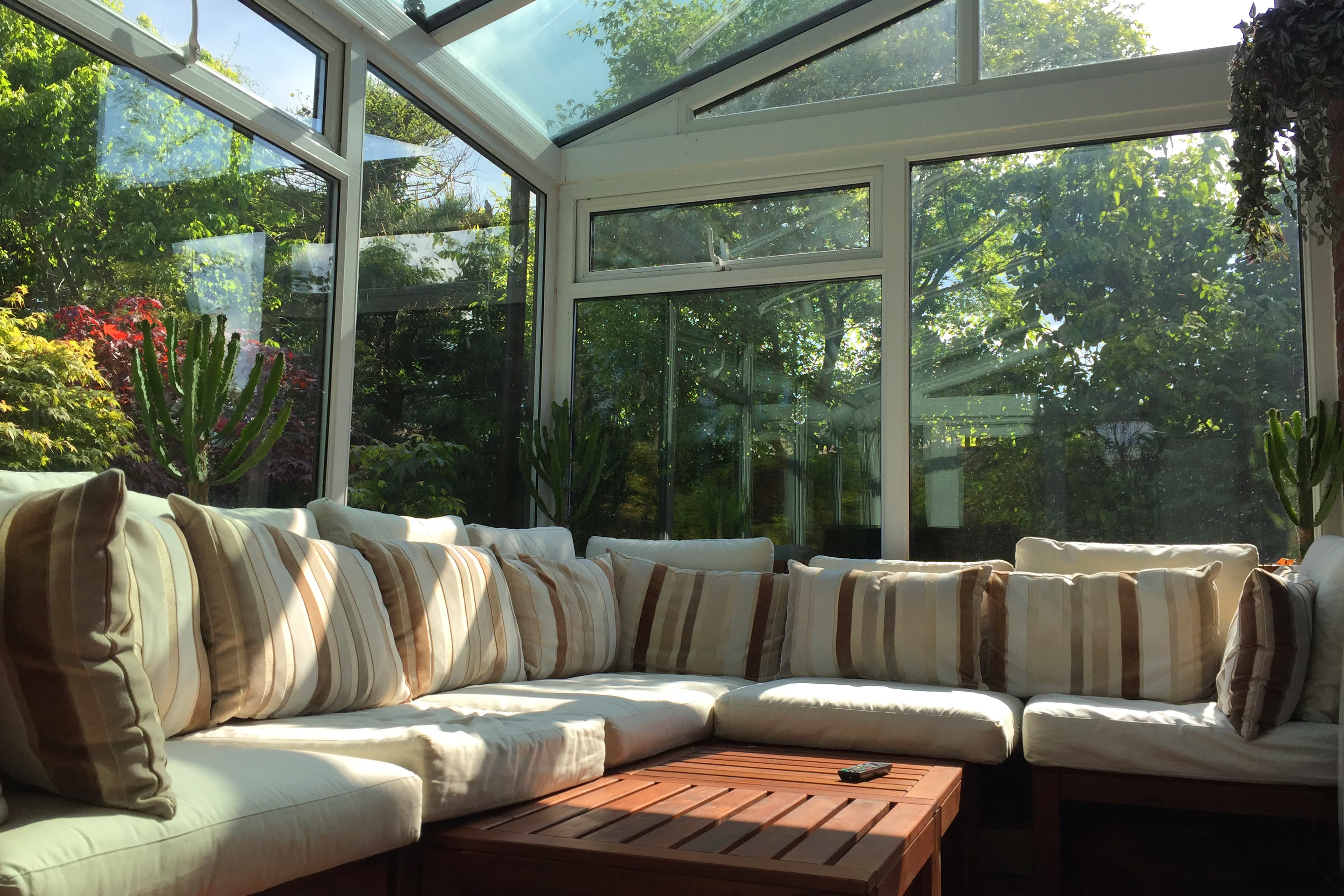 Wintergarten mit großem Ecksofa, Glasdach und Glaswänden, umgeben von Pflanzen und mit Blick auf den grünen Garten.