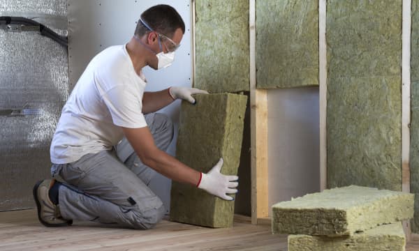 Handwerker mit Sichtschutzbrille und Atemmaske legt Steinwolle in Holzrahmen in einer Hauswand ein
