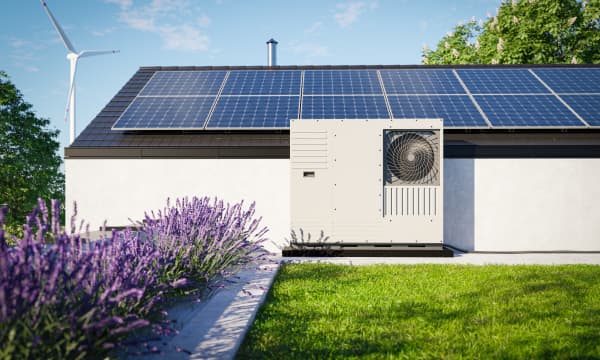 Wärmepumpe vor Haus mit Solaranlage auf dem Dach vor einem Lavendelbeet und grünem Rasen. Im Hintergrund steht ein Windrad