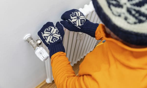 Kind in orangener Jacke und blauweißen Handschuhen und Mütze fasst auf Heizkörper