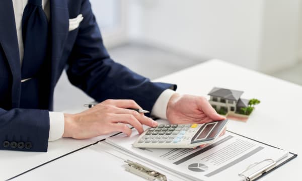 Mann in Anzug sitzt an einem Tisch mit Unterlagen und hält einen Taschenrechner in der Hand