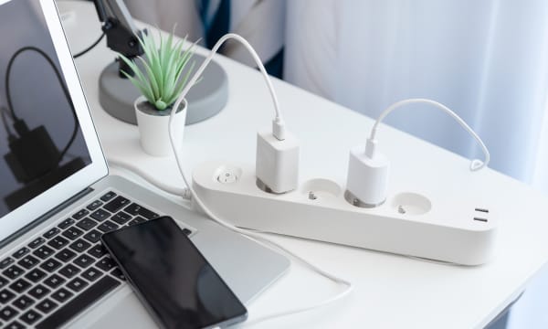 Weiße Steckerleiste mit eingesteckten Kabeln, die zu einem Laptop und einem Smartphone führen, die auf einem weißen Tisch liegen.