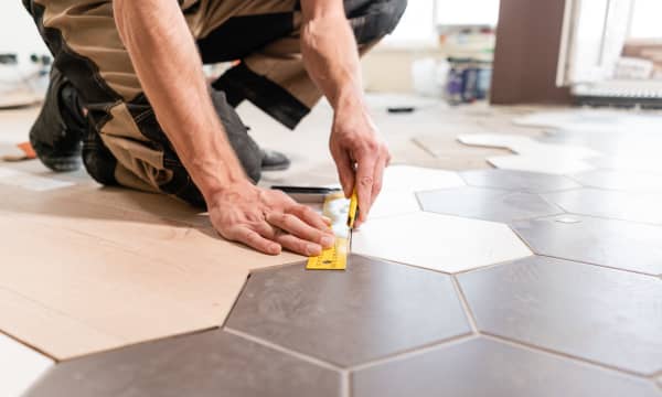 Mann in Arbeitskleidung kniet auf dem Boden und misst die Kante einer sechseckigen Bodenfliese zum Verlegen ab