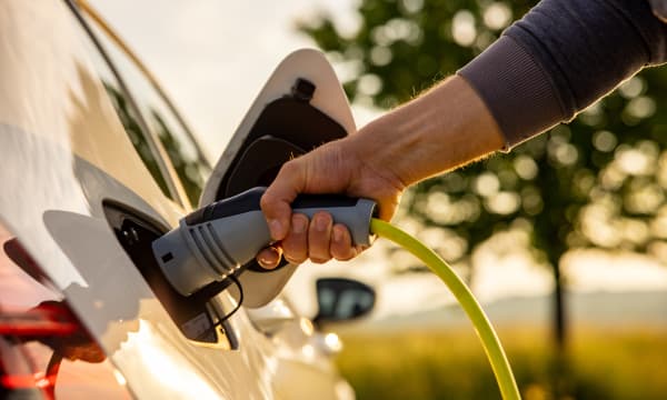 Ein Mann steckt einen Ladestecker an ein Elektroauto an, im Hintergrund ist eine verschwommene grüne Landschaft zu sehen