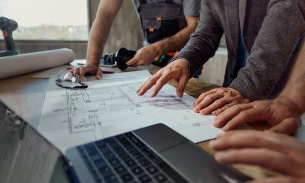 Drei Personen stehen an einem Tisch, auf dem Baupläne, ein Bohrer und ein Laptop liegen