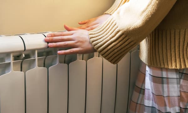 Frau in beigem Pullover und kariertem Rock wärmt ihre Hände an einem Heizkörper