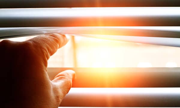 Hand einer Person, die die Lamellen einer Jalousie öffnet, dabei scheint die Sonne scheint durch das Fenster ins Hausinnere.
