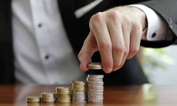 Mann in Hemd und Sakko stapelt Münzen auf ansteigend hohe Stapel auf einem Tisch