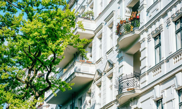 Seitliche Ansicht der weißen Fassade eines Mehrfamilienhauses in Berlin mit Stuck und Balkonen, davor ein grüner Baum