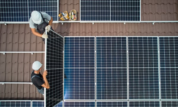 Ansicht von oben auf ein braun gedecktes Dach, auf dem zwei Arbeiter Solarmodule installieren