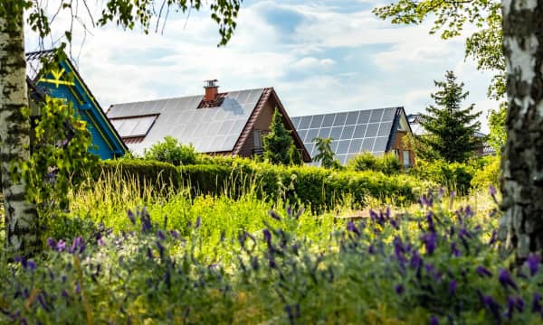 Mehrere Wohnhäuser mit Dächern, auf denen Solaranlagen installiert sind, mit grüner Wiese und Bäumen im Vordergrund unter blauem, leicht bewölktem Himmel