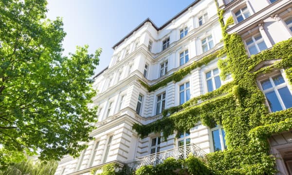 Blick von der Straße nach oben auf die weiße, teilweise mit Grünpflanzen bewachsene Fassade eines Mehrfamilienhauses