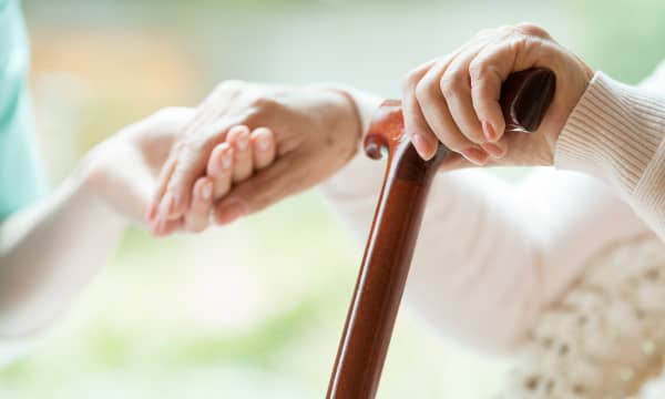 Nahaufnahme der Hände zweier Personen. Eine jüngere Person hält die Hand der älteren, die in der anderen Hand einen Gehstock hat