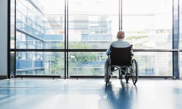 Senior im Rollstuhl blickt aus den bodentiefen Fenstern eines Gebäudes nach draußen