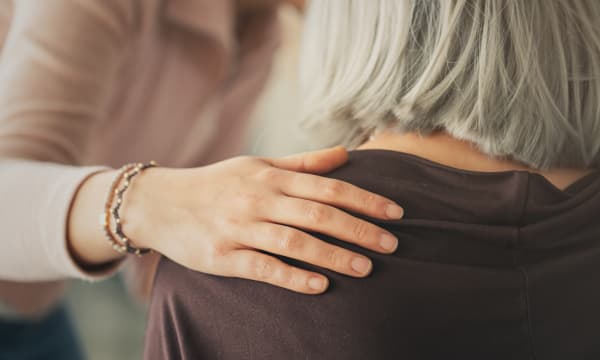 Jüngere Frau legt einer Seniorin mit kurzen grauen Haaren eine Hand auf die Schulter