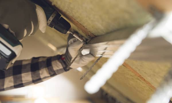 Nahaufnahme der Hände eines Arbeiters, der mit einem Akkuschrauber Bauteile innen an einer Dachschräge befestigt