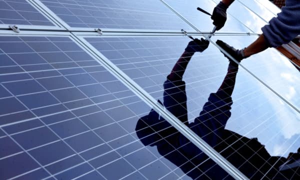 Blauschwarze Solarmodule werden montiert. Gespiegelt ist der Handwerker zu sehen, der eine Schraube festdreht.