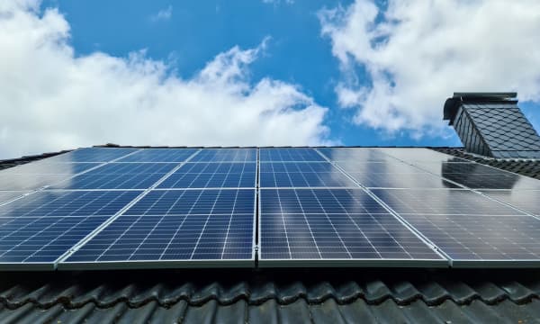 Blick von unten auf ein grau gedecktes Dach mit Schornstein und Solarmodulen bei blauem, leicht bewölktem Himmel