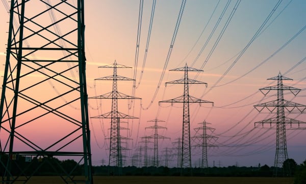 Mehrere Hochspannungsmasten in ländlicher Umgebung, die sich vor einem bewölkten Himmel bei Sonnenuntergang abzeichnen