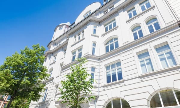 Saniertes Mehrfamilienhaus mit edler, weißer Fassade vor blauem Himmel mit grünen Bäumen davor