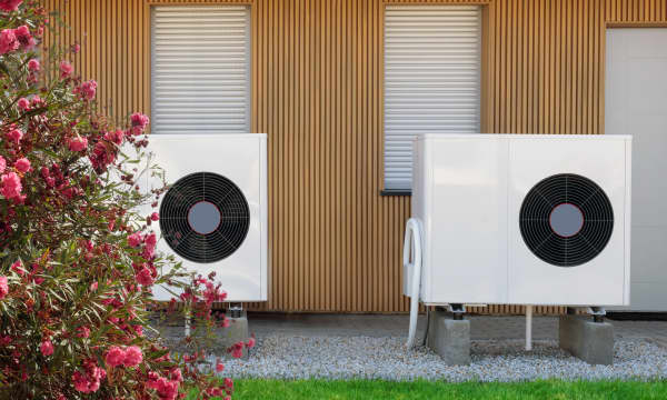 Zwei weiße Wärmepumpen vor einem Gebäude mit Holzfassade und mit Rollläden verschlossenen Fenstern