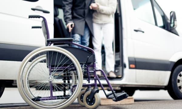 Rollstuhl steht vor einem weißen Van, aus dem ein Mann mit Gehstock aussteigt, wobei er von einer zweiten Person gestützt wird