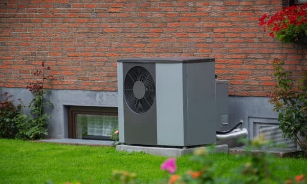 Graue Luftwärmepumpe vor der Ziegelfassade eines Hauses mit grüner Wiese im Vordergrund