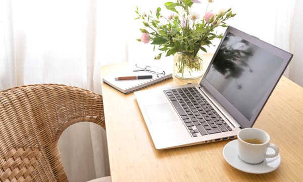 Tisch mit aufgeklapptem Laptop, Kaffeetasse, Notizblock und Blumenvase