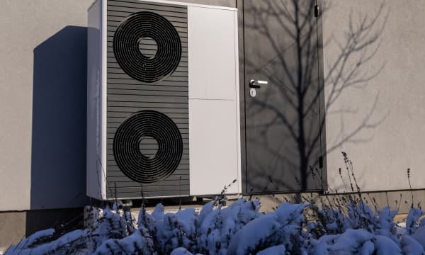 Luftwärmepumpen an der Fassade neben einer Eingangstür mit schneebedeckten Pflanzen im Vordergrund