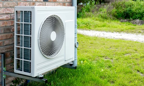 Luftwärmepumpe mit weißem Gehäuse an einer Hauswand mit Naturstein-Fassade mit grüner Wiese