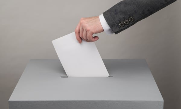 Mann mit Anzug steckt einen Zettel in eine Wahlurne, Aufnahme der Hand und des Arms