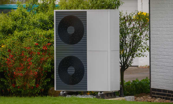 Luftwärmepumpe vor grüner Hecke an der Ecke eines Wohnhauses