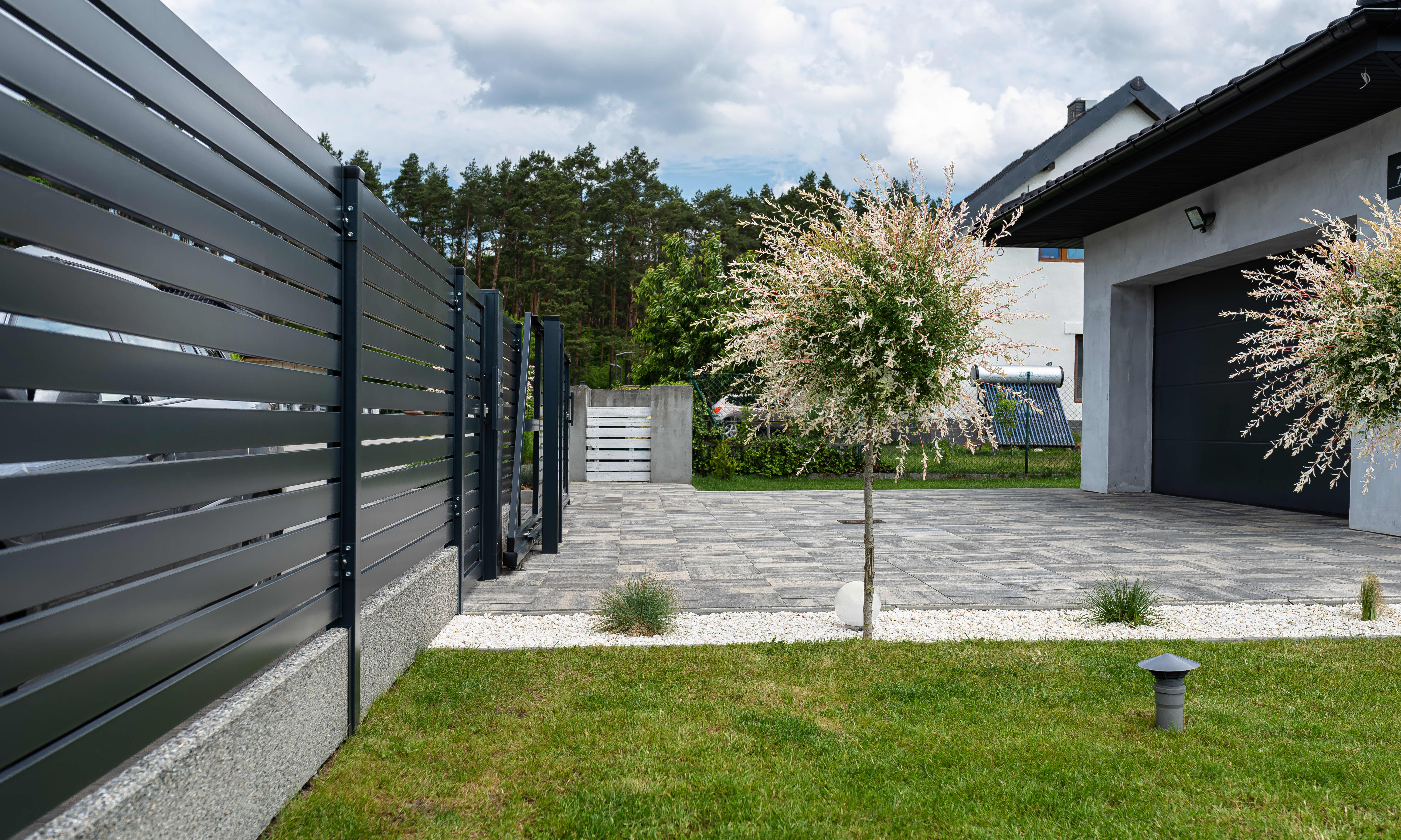 Modernes Wohnhaus mit gepflasterter Einfahrt und grüner Wiese. Das Grundstück wird durch einen hohen Zaun begrenzt.