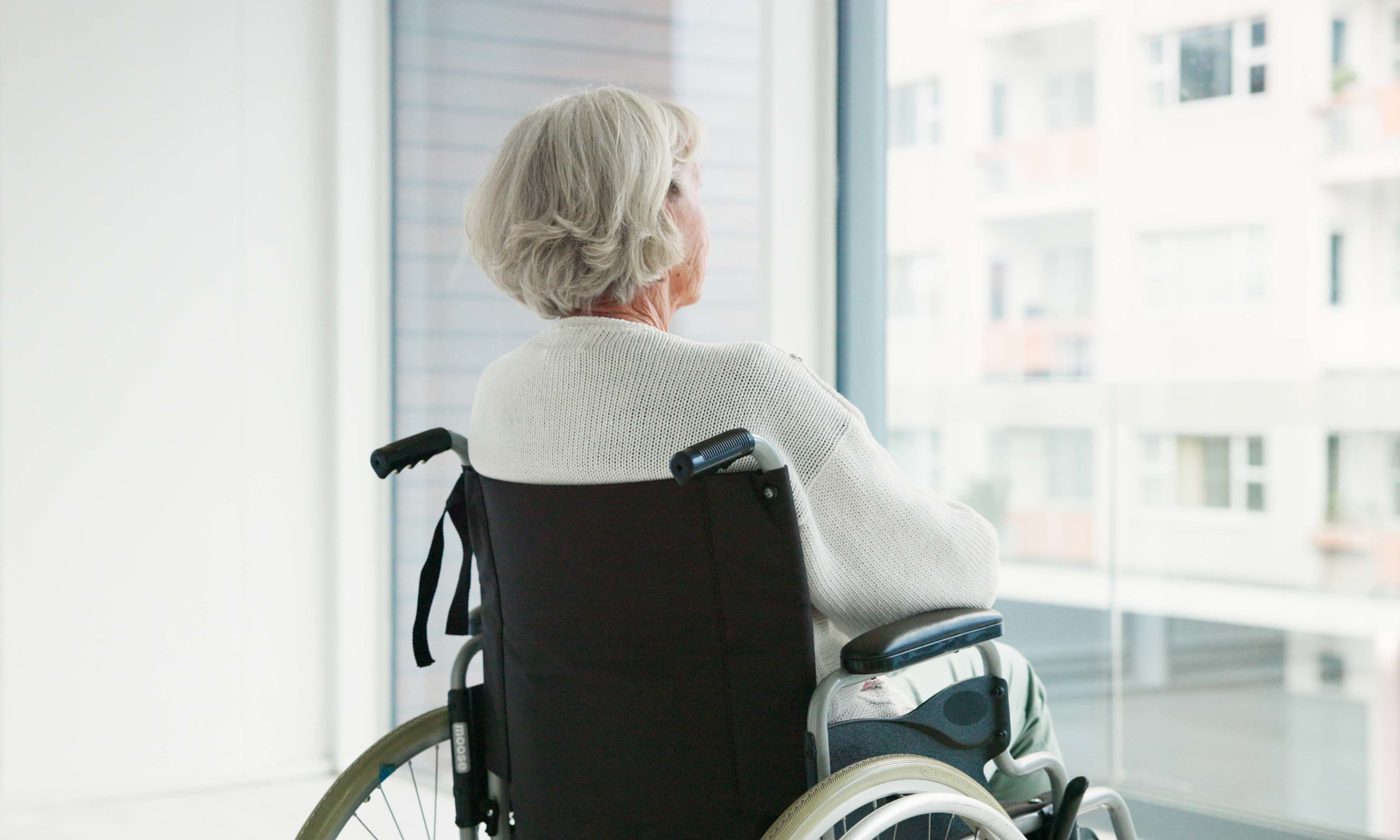 Blick auf eine Seniorin im Rollstuhl, die in einem hellen Raum aus dem Fenster blickt