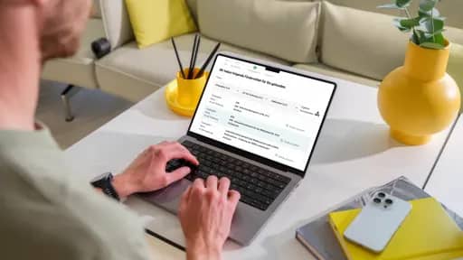Person arbeitet am Laptop auf einem weißen Tisch mit Smartphone und gelber Vase