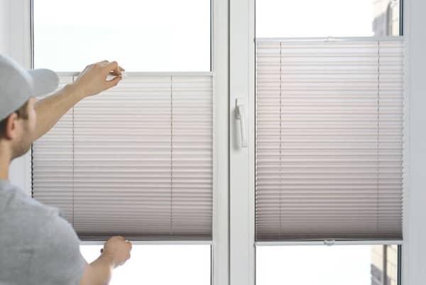 Plissee am Fenster einstellen – Mann reguliert helles Faltrollo an weißem Kunststofffenster