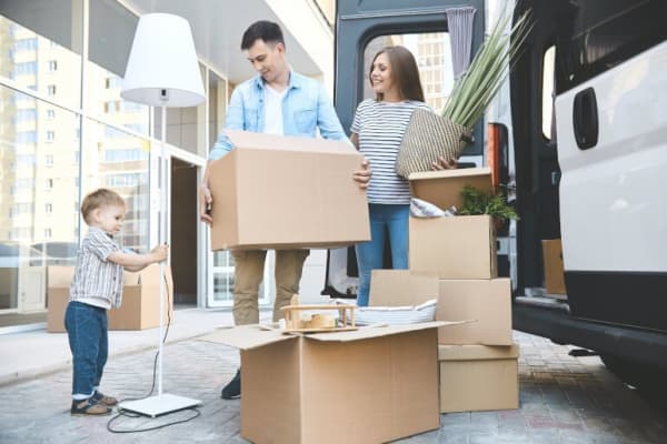 Bild zum Thema privater Umzug auf dem eine Familie mit Umzugskisten, einer Lampe und weiteren Gegenständen hinter einem Transporter steht