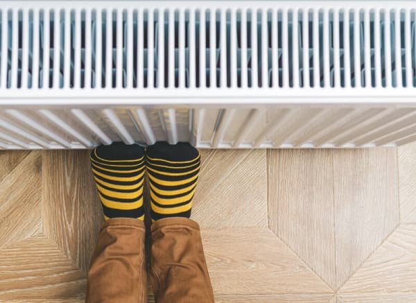 Person mit gelb-schwarz gestreiften Socken wärmt die Füße an einem eingeschalteten Heizkörper, während sie auf einem Holzfußboden sitzt.