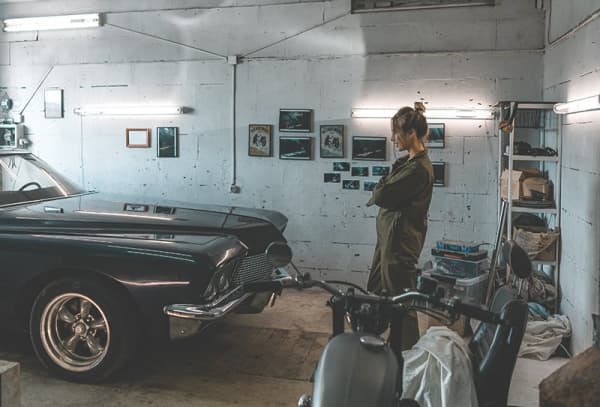 Eine junge Frau in olivfarbenem Overall steht im Inneren einer Garage und blickt auf einen schwarzen Oldtimer.