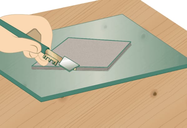 Hand führt einen Glasschneider an einer Schablone entlang, die auf einer Glasplatte liegt.