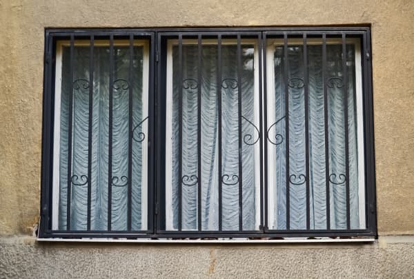Das Bild zeigt ein rechteckiges Fenster mit einem schwarzen Metallgitter. Hinter dem Gitter sind weiße Fensterrahmen und ein gemusterter, blauer Vorhang zu sehen. Die Hauswand besteht aus einer rauen, beigen Putzoberfläche.


Fenster mit Gitter zum Öffnen und Vorhang an einer Hausfassade
