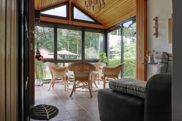 Wintergarten mit Holzdecke, großen Glasfenstern, Rattanmöbeln, Fliesenboden und Blick ins Grüne.