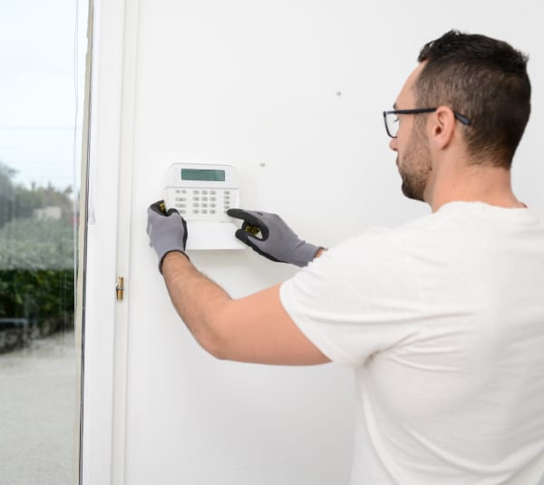 Ein Handwerker mit Handschuhen und Brille installiert eine weiße Alarmanlagen-Tastatur an einer Innenwand neben einer Glastür. Er hält das Gerät mit beiden Händen und deutet auf die Tasten; im Hintergrund ist draußen eine Gartenansicht zu erkennen.