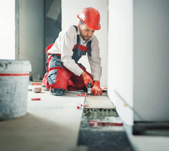 Ein Handwerker in roter Arbeitskleidung und Schutzhelm kniet auf einer Baustelle und verlegt großformatige Bodenfliesen. Er nutzt ein Werkzeug zum Ausrichten der Fliesen; Abstandshalter und ein Mörtelkübel sind im Vordergrund zu sehen. Heller Innenraum mit noch unfertigen Wänden.