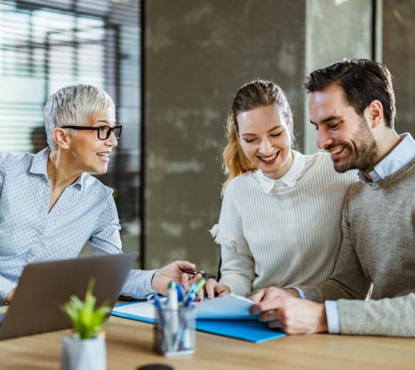 Innenaufnahme eines modernen Büros: Eine ältere Person mit Kurzhaarfrisur erklärt etwas und zeigt auf Dokumente, zwei jüngere Personen sitzen daneben und hören zu. Auf dem Holztisch liegen Ordner, Stifte und ein Notizbuch, daneben steht ein geöffneter Laptop und eine kleine Topfpflanze. Licht fällt durch große Fenster mit Lamellen in den Raum.