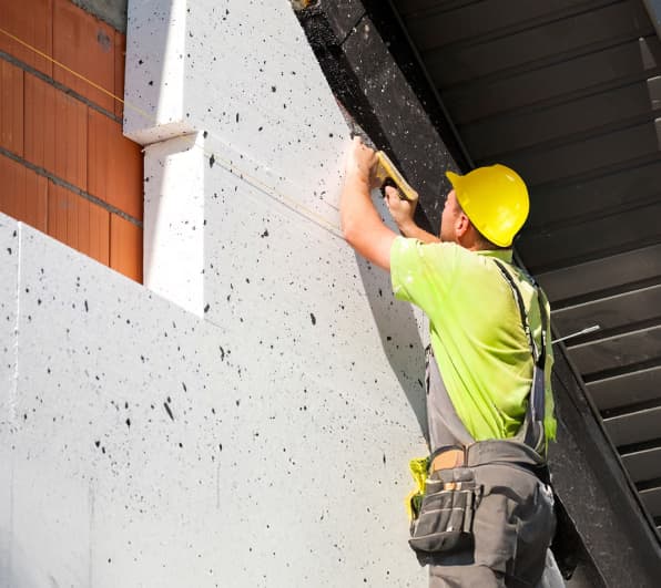 Nahaufnahme einer Baustelle: Ein Handwerker mit gelbem Schutzhelm richtet weiße, gesprenkelte EPS-Dämmplatten an einer Ziegelaußenwand aus und schneidet die Kante unter einem Dachüberstand zu. Sichtbar sind Mauerwerk, Dämmplatten, Schnur zum Ausrichten und Werkzeuge.