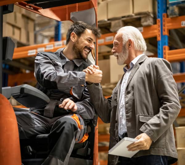 Zwei Personen in einem Lagerhaus: ein sitzender Gabelstaplerfahrer in Arbeitskleidung und ein älterer Mann mit Notizblock geben sich die Hand. Im Hintergrund sind hohe Regale mit Kartons zu sehen, Teile des Gabelstaplers sind sichtbar. Gesichtszüge sind unkenntlich gemacht.
