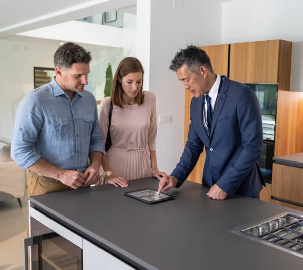 In einer modernen Einbauküche stehen drei Personen an einer großen Kochinsel. Ein Berater im Anzug zeigt auf ein Tablet, während ein Paar in Freizeitkleidung aufmerksam zusieht. Sichtbar sind dunkle Arbeitsplatte, integriertes Kochfeld und hohe Holzschränke im Hintergrund.
