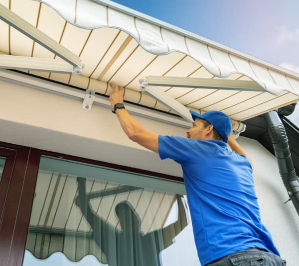 Tageslichtaufnahme an einer Hausfassade: Ein Monteur in blauem Shirt und Kappe arbeitet über einer großen Glas-Schiebetür an einer ausgefahrenen, hellen Gelenkarmmarkise mit Wellenvolant. Sichtbar sind die Metallarme, die Wandhalterungen und die Regenrinne am Dachrand; im Glas spiegelt sich der Arbeitsbereich.