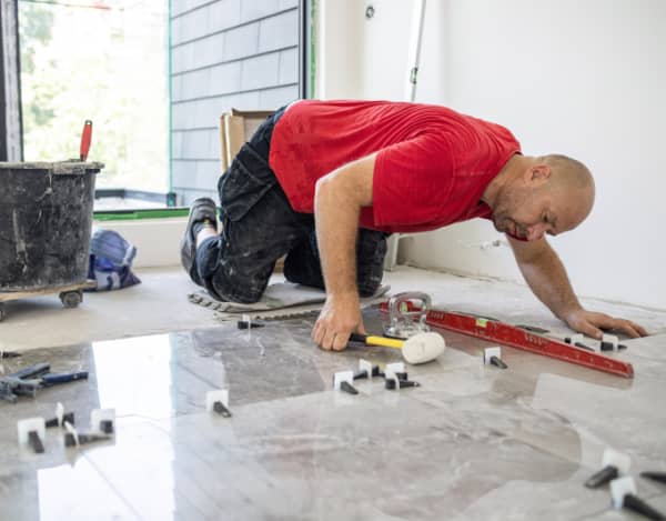 Ein Handwerker im roten T‑Shirt kniet in einem hellen Raum und richtet eine großformatige, glänzende Bodenfliese aus. Vor ihm liegen Wasserwaage, Gummihammer, Saugheber und Fliesen-Nivellierkeile; daneben ein Mörtelkübel und weitere Werkzeuge. Im Hintergrund eine offene Fensteröffnung und unverputzte Wände.